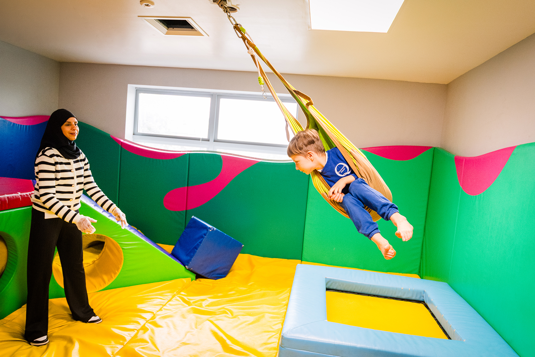 A boy playing in a swing in a soft play area. He is being supervised by a teacher.