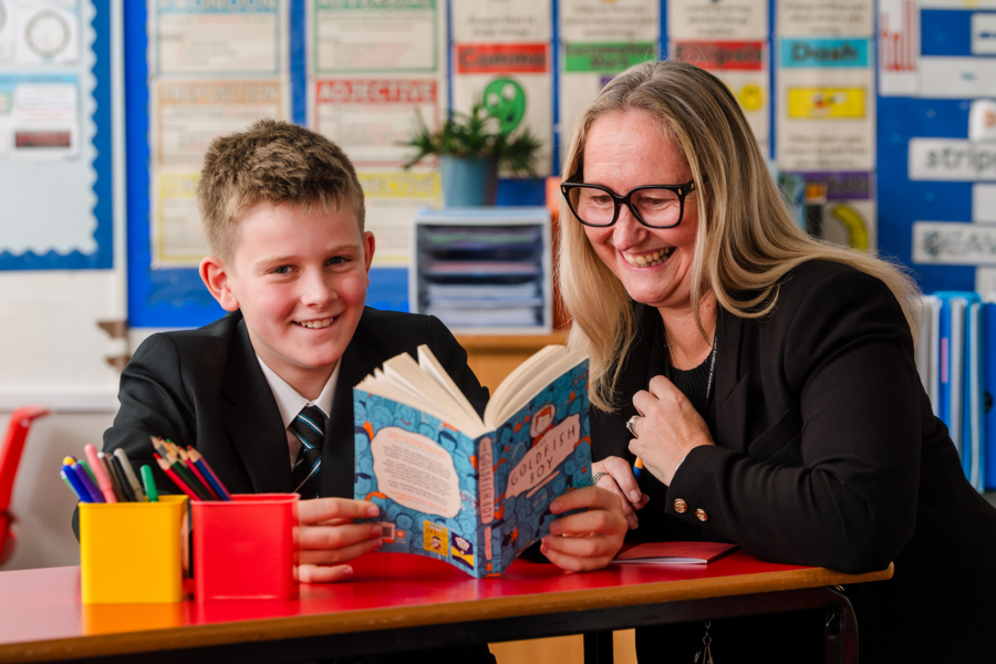 Photograph of a teacher and student in a classroom setting. The student is reading from a book.